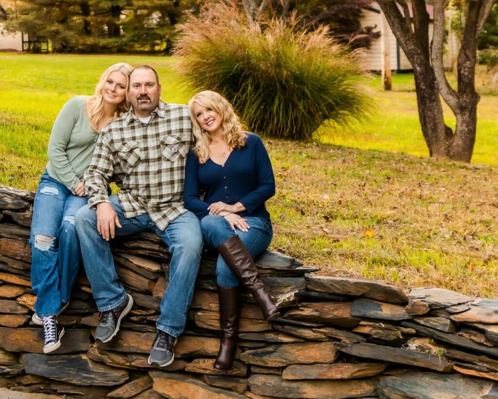 Three people pose smiling on a stone wall. Autumn colors fill the background. They wear casual, comfortable clothing and seem relaxed and happy together.