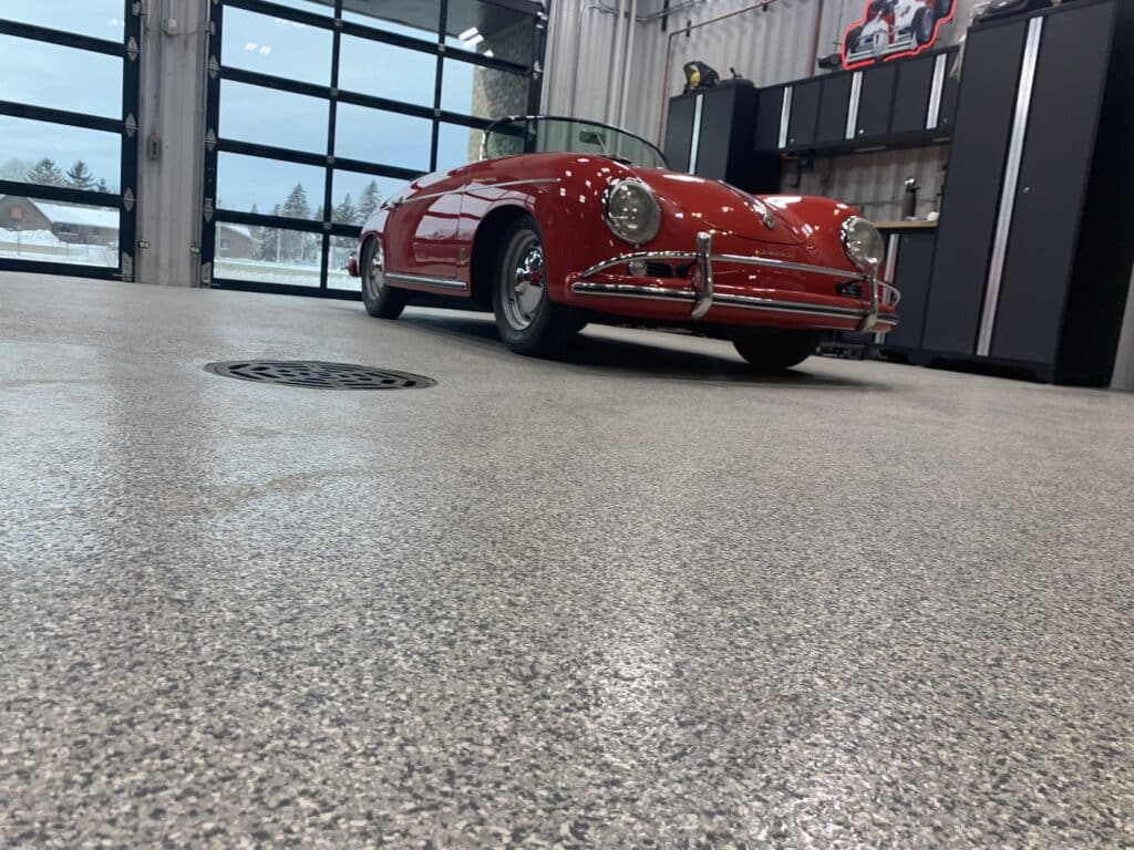 A red vintage convertible car is parked inside a clean garage with large windows, a concrete floor, and storage cabinets along the wall.