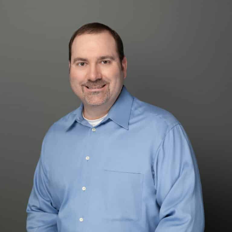 A smiling person wearing a blue button-up shirt poses against a plain grey backdrop for a professional headshot. They appear friendly and approachable.