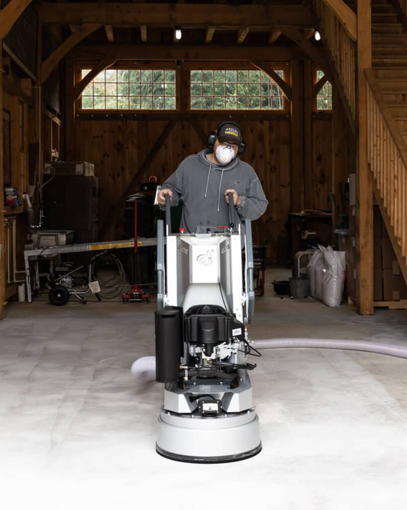 A person in protective gear operates a large floor polishing machine inside a wooden workspace with tools and equipment visible in the background.