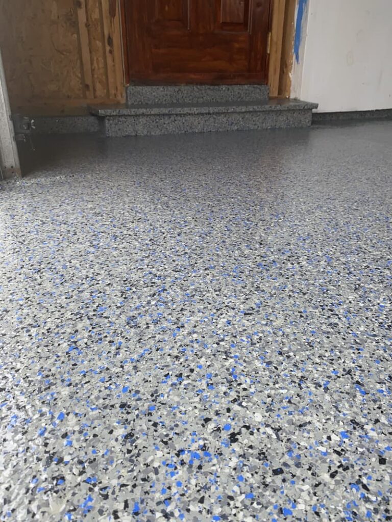 The image shows a speckled epoxy floor with blue, gray, and white chips. There's a wooden door and steps leading to it in the background.