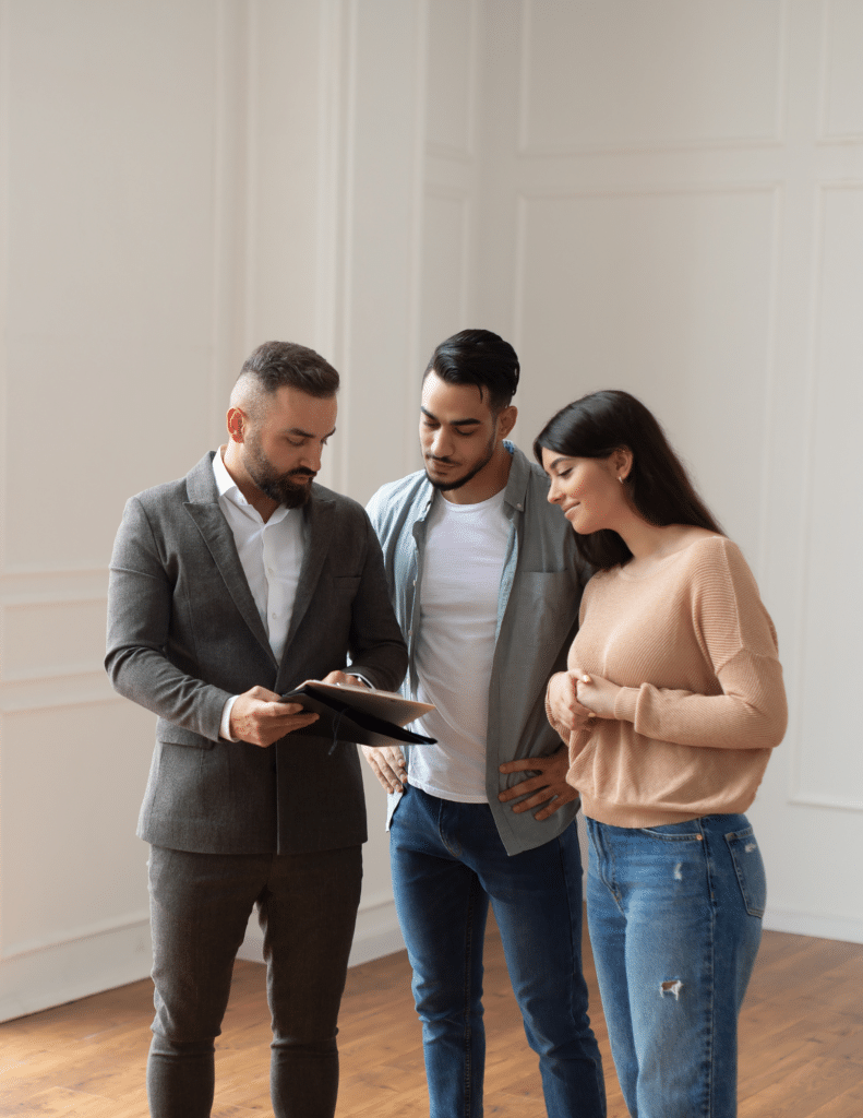 Three people are standing indoors, one holding a document. Two are attentively looking at the paper. They appear engaged in a serious discussion.